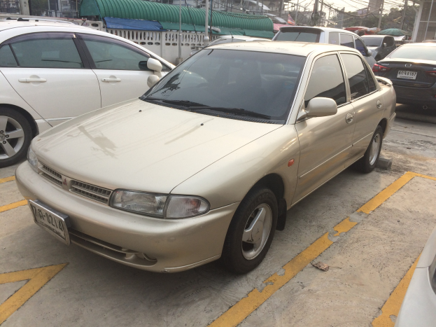 Goldfarbener Wagen auf einem Parkplatz neben anderen Fahrzeugen geparkt, umgeben von Gebäuden, Strommasten, Drähten, Bäumen und einem klaren blauen Himmel.