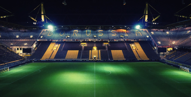 Ein Fubballfeld nachts, erleuchtet von hellen Stadionlichtern, mit einigen Menschen auf den R├Ąngen und saftig gr├╝nem Rasen.