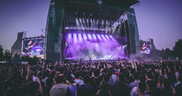 Eine große Menschenmenge bei einem Musikfestival schaut auf einer Bühne mit Instrumenten, Lichtern und Bildschirmen, die vor einem Gebäude, Bäumen und einem blauen Himmel aufgebaut sind.