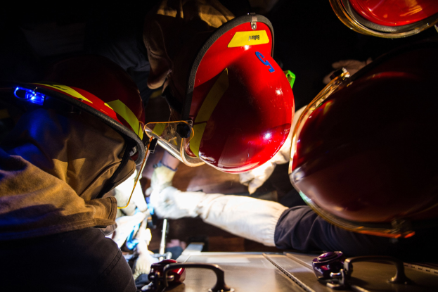 Feuerwehrleute in Helmen und Handschuhen arbeiten an einem Feuerwehrauto in einem schwach beleuchteten Bereich mit einem Tisch voller Vorräte auf der rechten Seite.