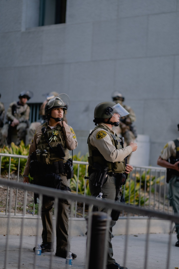 Gruppe von Polizisten in Uniform mit Helmen und Stiefeln vor einem Gebäude stehend, einige halten Waffen, mit einem Metallzaun im Vordergrund und Pflanzen und einer Wand im Hintergrund.