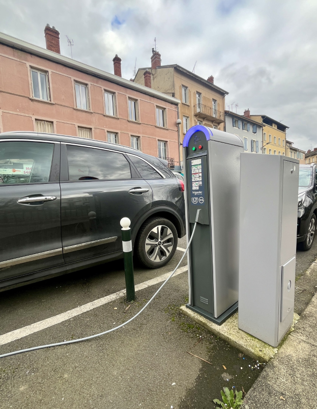 Elektroauto-Laden an einer stra√enseitigen Station mit Gebäuden, Laternenmästen und bewölktem Himmel im Hintergrund.