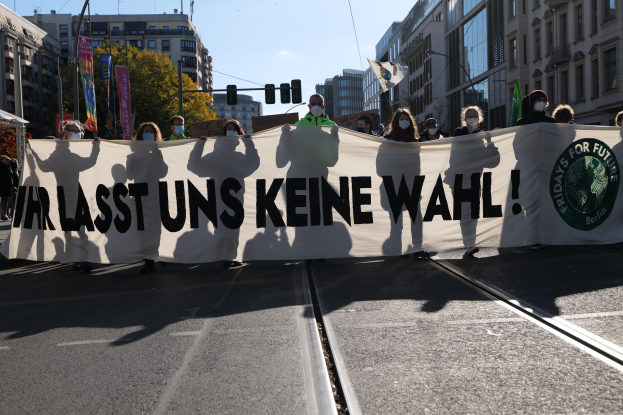Eine Gruppe von Menschen mit Masken hält ein Transparent mit der Aufschrift "Wir sind die letzten keine wahl" vor Gebäuden, Bäumen, Laternenmasten, Ampeln und einem Zelt, mit dem Himmel im Hintergrund.