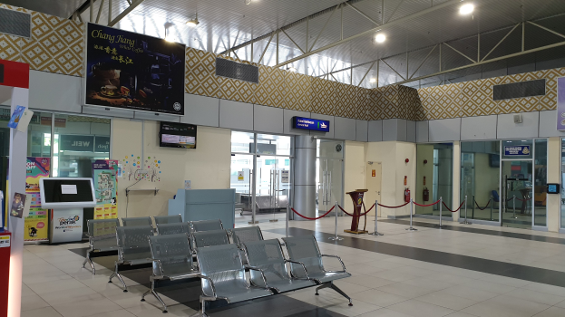 Leerer Wartebereich am Flughafen mit Stühlen, Absperrpoller mit Seilen, Informationsschildern, Wandbildschirmen, Deckenleuchten und Glastüren.