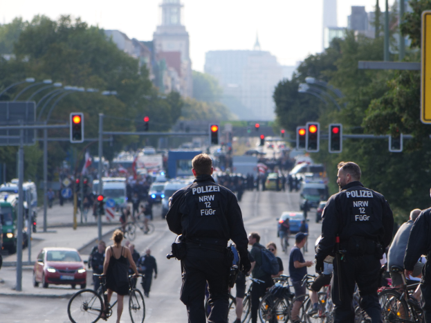 Polizeibeamte mit Fahrr√°dern auf einer von Bäumen gesäumten Stra√če mit Geb√°uden und einem klaren blauen Himmel im Hintergrund.