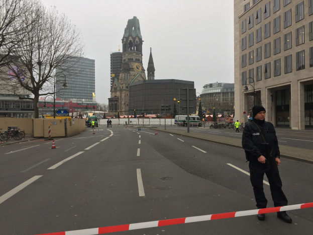 Ein Polizist in schwarzer Uniform und Mütze steht in einer Berliner Straße umgeben von Verkehrskegeln, Fahrrädern, Laternenmasten, Bäumen und Gebäuden, mit einem Kirchturm im Hintergrund bei klarem blauem Himmel.