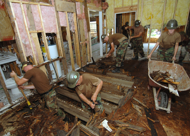 Eine Gruppe von Männern in Helmen, die Werkzeuge halten, arbeitet in einem Raum im Bau mit verstreuten Materialien und einem mit Holzstücken beladenen Wagen.