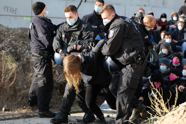 Gruppe maskierter Menschen in Kapuzenpullis, einige mit Waffen, umringt eine Frau, die von der Polizei eskortiert wird, mit Pflanzen, Steinen und einer Wand mit Protesttext im Hintergrund.