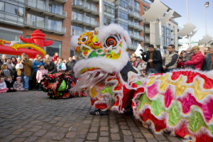 Vibrantes chinesisches Neujahrsfest in Amsterdam mit einer Löwen-Tanzvorstellung und einer Zuschauermenge, einige halten Kameras, vor einem Hintergrund aus Gebäuden, Laternenmasten und einem klaren blauen Himmel.
