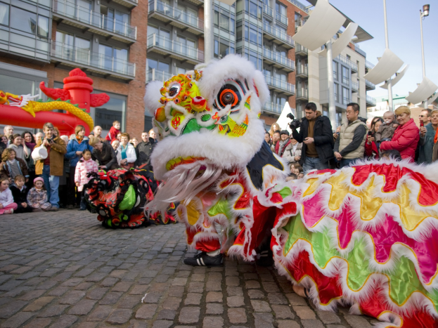 Vibrantes chinesisches Neujahrsfest in Amsterdam mit einer Löwen-Tanzvorstellung und einer Zuschauermenge, einige halten Kameras, vor einem Hintergrund aus Gebäuden, Laternenmasten und einem klaren blauen Himmel.