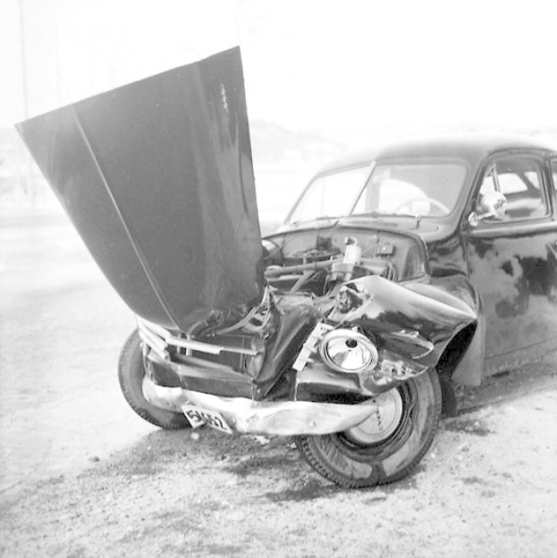 Schwarzes und weißes Foto eines schwer beschädigten Autos am Straßenrand mit eingedellter Motorhaube, zersplitterten Fenstern und offener Fahrertür, die den Motor zeigt.