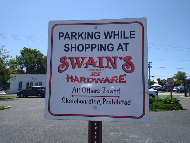 Ein Parkplatz mit einem Schild, auf dem "Parken beim Einkaufen bei Swain's Hardware" steht, im Vordergrund, mit Fahrzeugen, Pflanzen, Bäumen, Gebäuden, Polen, Drähten und einem klaren blauen Himmel im Hintergrund.