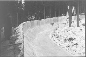 Ein Schwarz-Weiß-Foto von einer Person, die auf einem Bobschlitten eine schneebedeckte Bahn hinunterfährt, umgeben von einem Zaun und Bäumen, mit ein paar Menschen im Hintergrund.