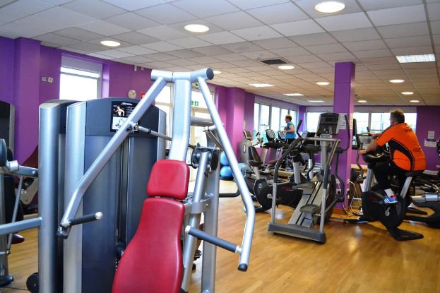 Ein Fitnessstudio mit mehreren Menschen, die an verschiedenen Geräten trainieren, mit einer Decke und Lampen oben im Bild.