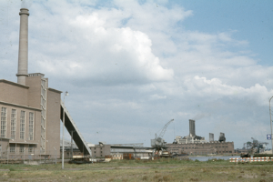 Ein altes Foto einer Fabrik mit Rauch, der aus ihren Schornsteinen aufsteigt, umgeben von verschiedenen Industriegebäuden, mit grasbewachsenem Boden und einem bewölkten Himmel.