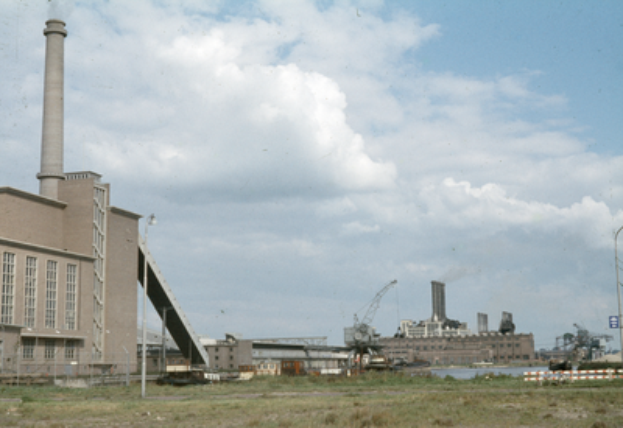 Ein altes Foto einer Fabrik mit Rauch, der aus ihren Schornsteinen aufsteigt, umgeben von verschiedenen Industriegebäuden, mit grasbewachsenem Boden und einem bewölkten Himmel.