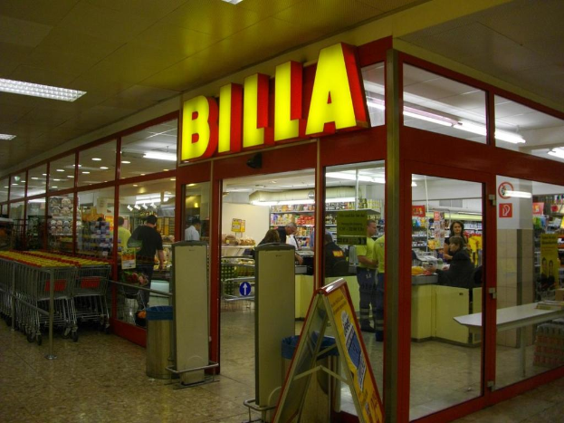 Ein Markt mit einem Eingang in der Mitte, Mülltonnen daneben, einer Reihe von Einkaufswagen auf der linken Seite, Deckenbeleuchtung und einigen Menschen, die Produkte anschauen.