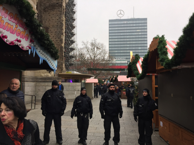 Polizeibeamte in schwarzen Uniformen stehen vor einem geschmückten Weihnachtsmarkt in Berlin, mit festlichen Ständen, Gebäuden, Bäumen und einem klaren blauen Himmel im Hintergrund.