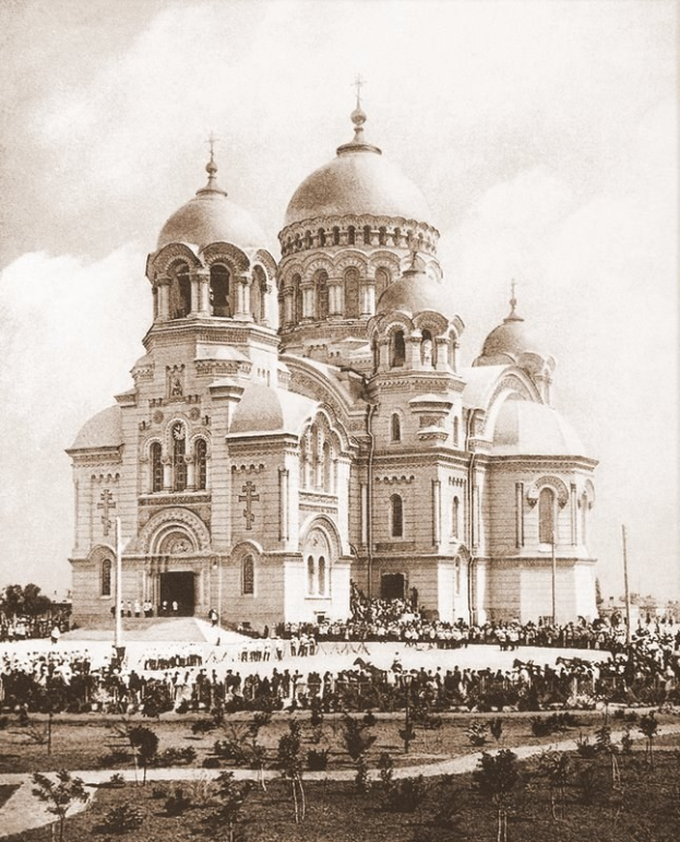 Schwarz-weiß-Fotografie der Erlöserkirche auf dem Blutsplatz in St. Petersburg, Russland, mit Menschen drumherum, Bäumen und Pfählen im Vordergrund und einem klaren Himmel im Hintergrund.