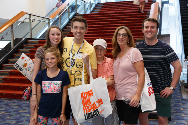 Eine Gruppe lachender Menschen mit Taschen steht zusammen vor einer Rolltreppe auf der Nationalen Buchmesse 2019, mit Treppen, Geländern und einer Wand im Hintergrund.