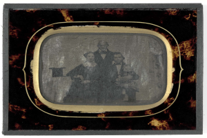 Ein Schwarz-Weiß-Foto eines Mannes im Anzug und einer Frau in unbekannter Kleidung, die vor einem alten Fernseher stehen und ernst schauen.