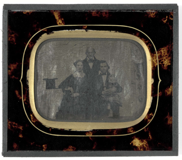 Ein Schwarz-Weiß-Foto eines Mannes im Anzug und einer Frau in unbekannter Kleidung, die vor einem alten Fernseher stehen und ernst schauen.