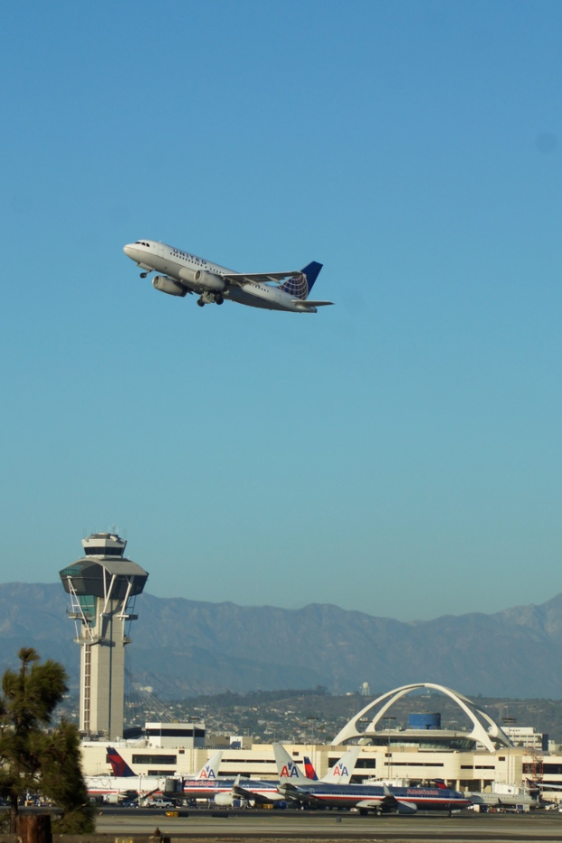 Flugplatzansicht mit weißer Kontrollturm, geparkten Flugzeugen, entfernten Gebäuden und einem Flugzeug am Himmel