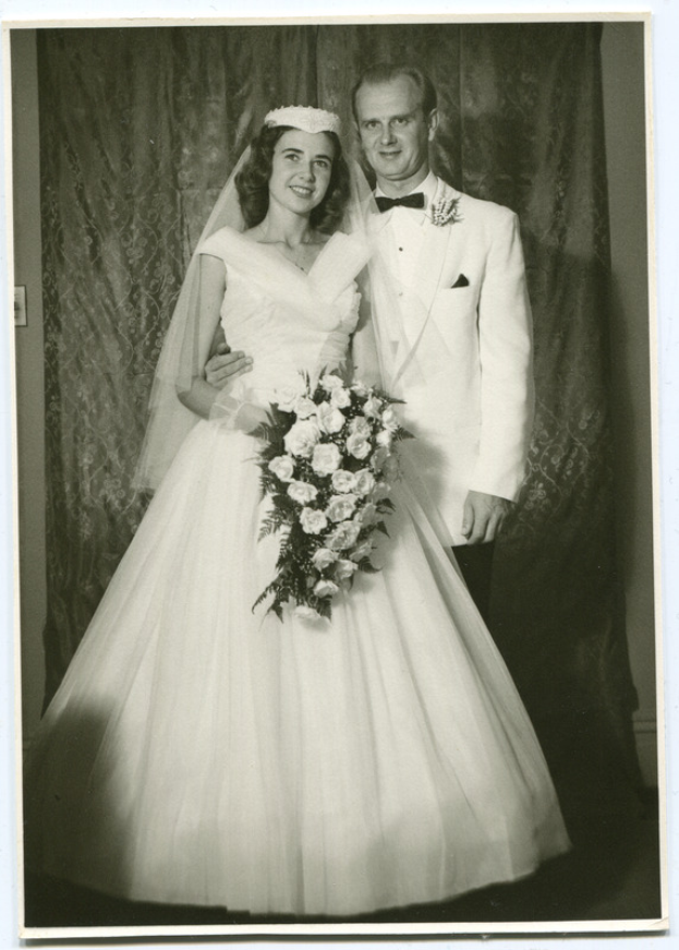 Ein Schwarz-Weiß-Foto einer Braut in einem weißen Kleid, die einen Strauß hält und von einem strahlenden Bräutigam begleitet wird, mit einem Vorhang im Hintergrund.