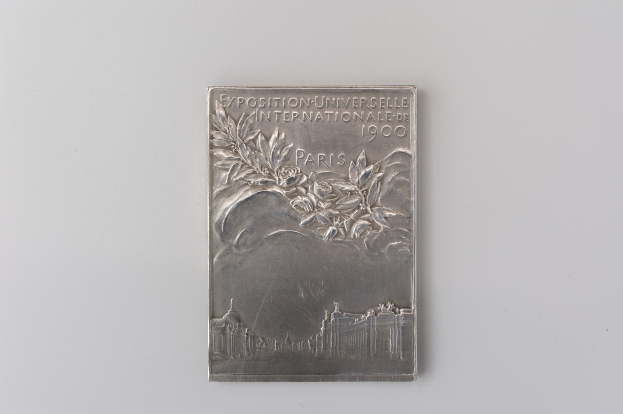 Silberne Plakette auf weißem Hintergrund mit der Aufschrift "Exposition Universelle Internationale de Paris 1900" und einem detaillierten Design.