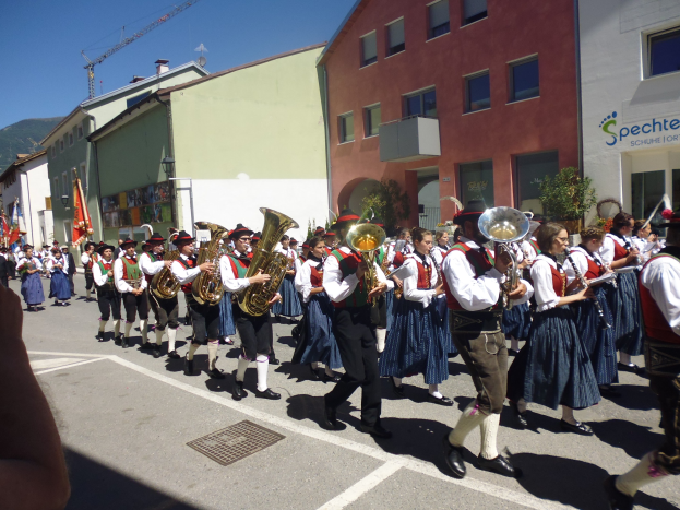 Eine Gruppe von Menschen in traditioneller bayrischer Tracht, die auf der Straße musizieren und dabei durch eine Straße mit Gebäuden, einigen Fahnen haltenden Personen und einem Hügel sowie einem blauen Himmel gehen.
