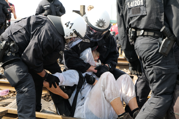Eine Gruppe von Polizisten in Einsatzausrüstung nimmt einen Mann in einem weißen Kleid auf einem Gleis fest, während andere im Hintergrund zu sehen sind.