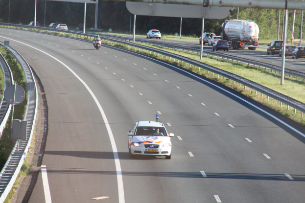 Ein Auto fährt auf einer Autobahn mit einem Polizeiwagen am Straßenrand, anderen Fahrzeugen, Geländern, Polen, Planken und Bäumen im Hintergrund.