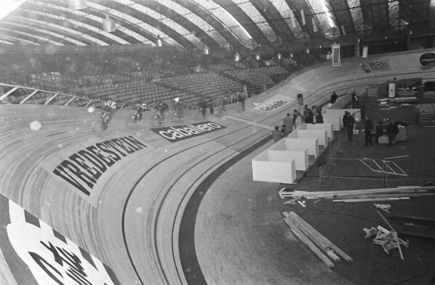 Ein Schwarz-Weiß-Foto eines Hallenstadions mit einer zentralen Laufbahn, ein paar Menschen auf der rechten Seite, Stühle auf der linken Seite und ein beleuchtetes Dach oben.