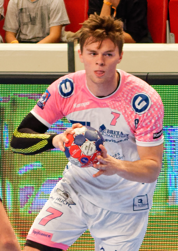 Ein Mann in einem pinken Hemd mit einem Fußball in der Hand, daneben ist eine andere Person Hand zu sehen und im Hintergrund sitzen ein paar Leute auf Stühlen.
