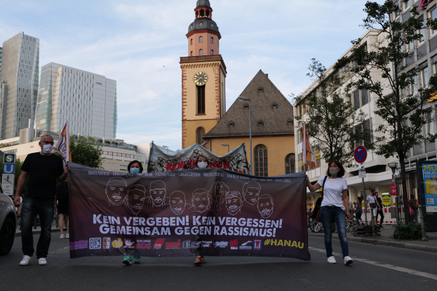Eine Gruppe von Menschen in Masken, die eine Straße entlanggehen und ein Banner halten, mit einem geparkten Auto links, Gebäuden und Bäumen im Hintergrund und einem klaren blauen Himmel darüber.