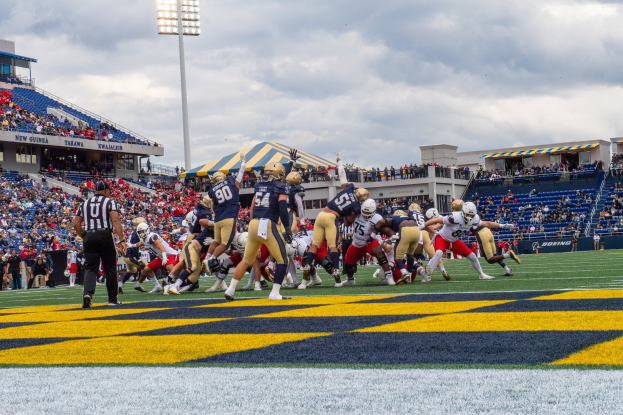 Fußballspieler mit Helmen und Sportbekleidung konkurrieren auf einem Feld mit Zuschauern, Stadionstrukturen und Text unten.