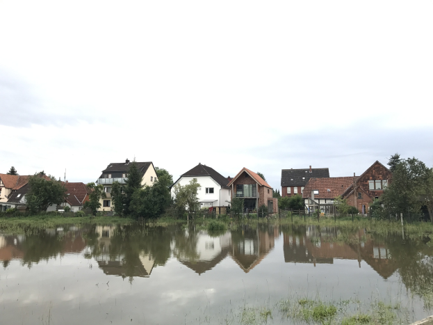 Eine überflutete Straße mit teilweise unter Wasser stehenden Häusern, umgeben von Gras, Pflanzen, Bäumen und Strommasten, unter einem sichtbaren Himmel.