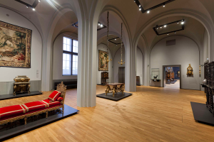 Ein geräumiger Raum im Kunsthistorischen Museum in Wien, Österreich, mit einer Couch mit Kissen, Tischen mit verschiedenen Gegenständen, Säulen, Fenstern und Deckenleuchten, sowie zahlreichen paintings an den Wänden.