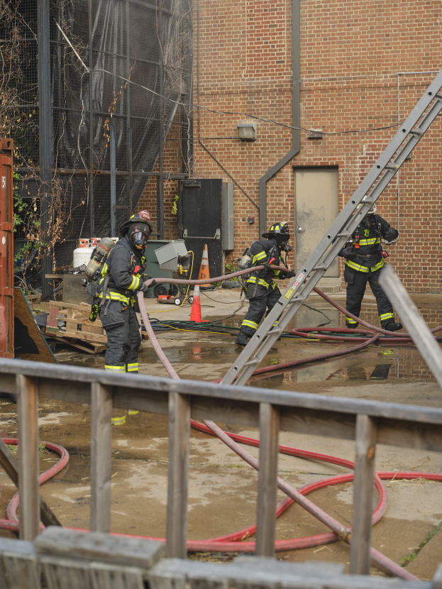Eine Gruppe von Feuerwehrleuten in Helmen, die Rohre halten, arbeitet daran, ein Gebäude Feuer zu löschen, mit verschiedenen Ausrüstungen, Strukturen und einem Baum in der Szene.