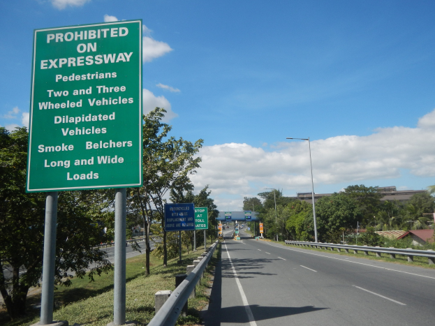 Grüne Highway-Schilder mit der Aufschrift "Verboten auf der Autobahn" mit Fahrzeugen, Laternenpfählen, Bäumen, Gebäuden und wolkenverhangenem Himmel im Hintergrund.