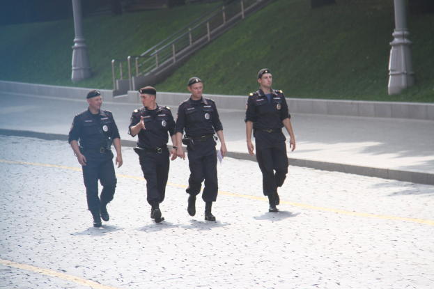 Drei Polizeibeamte in Uniform gehen öffentlich mit Stäben, Geländern, Treppen und Gras im Hintergrund.