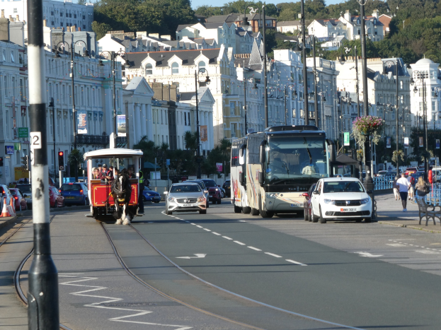 Eine belebte Stadtstraße mit hohen Gebäuden, Fahrzeugen, Fußgängern, Verkehrsampeln, Laternenpfählen, Bäumen und einem klaren blauen Himmel.