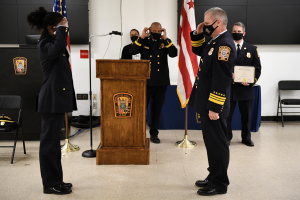 Gruppe uniformierter Personen in Masken auf einem Podium mit einem Mikrofon, umgeben von Fahnen und Möbeln, während einer Vereidungszeremonie, bei der eine Person ein Zertifikat hält.