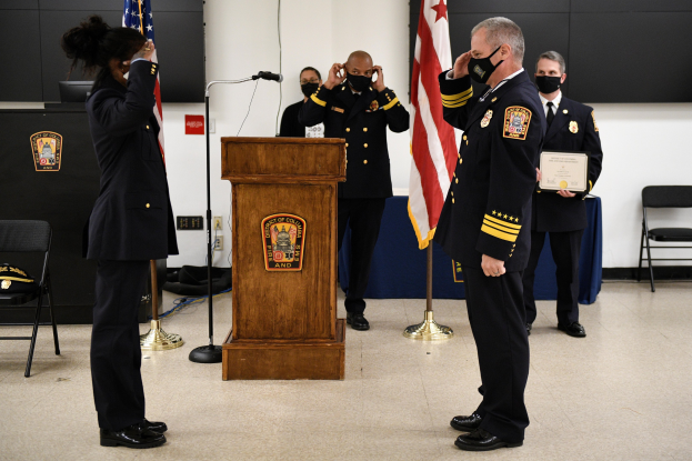 Gruppe uniformierter Personen in Masken auf einem Podium mit einem Mikrofon, umgeben von Fahnen und Möbeln, während einer Vereidungszeremonie, bei der eine Person ein Zertifikat hält.