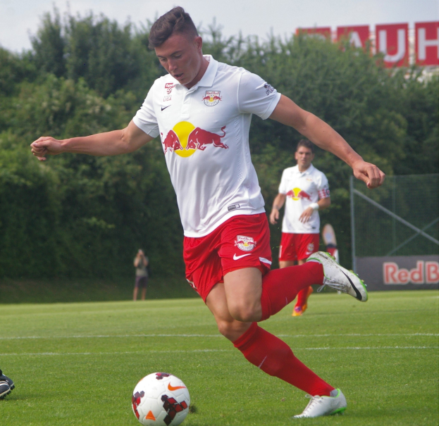 Ein professioneller Fussballspieler tritt einen Ball auf einem grünen Feld mit Bäumen und einem klaren blauen Himmel im Hintergrund, mit "RB Leipzig" auf einer Tafel dahinter.