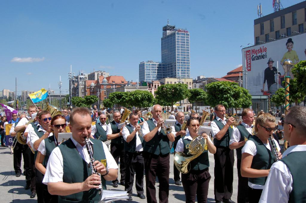 Eine Gruppe von Menschen in einheitlichen Kostümen, die verschiedene Musikinstrumente im Freien spielen, mit Bäumen, Pfählen und einem Banner mit Text und Bildern im Hintergrund.