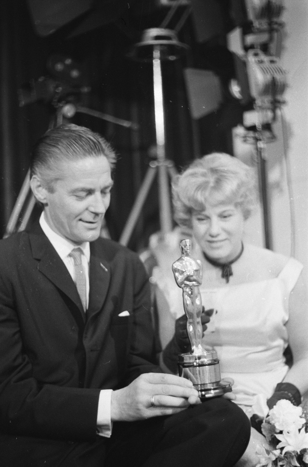 Ein Mann in einem schwarzen Blazer, weißem Hemd und Krawatte hält eine Oscar-Statue in der Hand und sitzt neben einer Frau, die einen Blumenstrauß hält, beide in festlicher Kleidung, mit Licht im Hintergrund, in Schwarz-Weiß.
