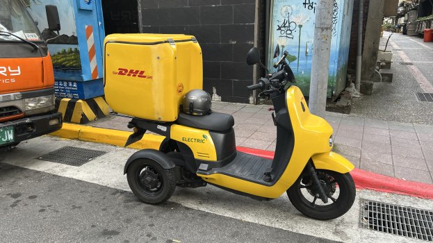 Ein gelber Elektro-Scooter mit Helm steht auf einer Straße neben einem Lastwagen, mit Gebäuden und Passanten im Hintergrund.