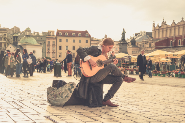 Ein Mann sitzt auf dem Boden und spielt eine Akustikgitarre in einem belebten Stadtplatz, umgeben von Passanten, mit einer Tasche auf dem Gehweg und Gebäuden, einer Statue und einem klaren Himmel im Hintergrund.