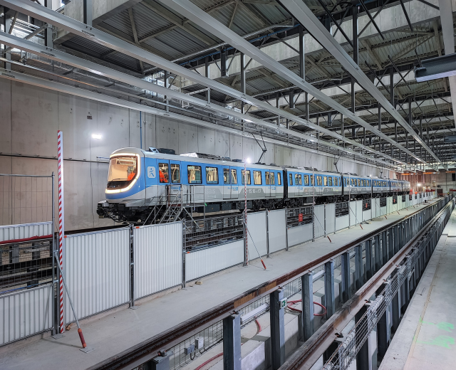 Blauer und weißer Zug fährt durch eine große Indoor-Station mit einer Plattform auf der rechten Seite und einer Wand im Hintergrund, mit einer Decke mit Stangen und Lampen.